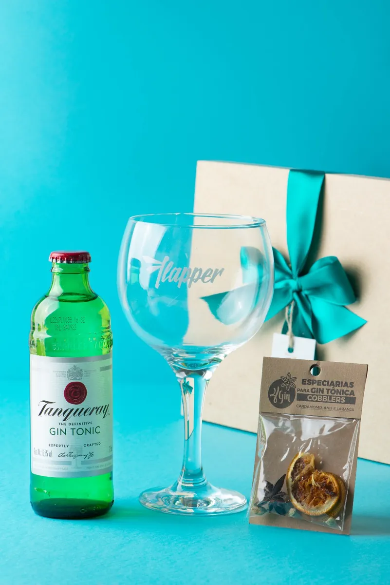 Taça de gin gravada, tônica e especiarias sobre fundo turquesa